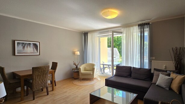 Una sala de estar moderna con una mesa de centro de vidrio, un sofá, una mesa de comedor con sillas y vistas al exterior a través de amplios ventanales.