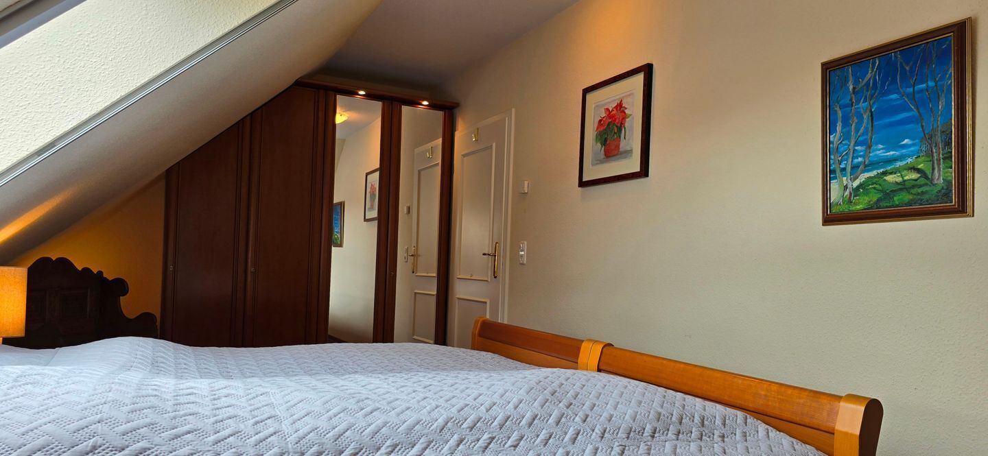 A bedroom with a sloped ceiling, a bed with a white quilt, a wooden headboard, a painting of trees, and a door leading to another room.