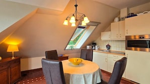 Un espace repas comprenant une table, des chaises, une lampe, une cuisine avec des placards et des appareils électroménagers, et un plafond en pente doté d’un puits de lumière.