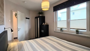 Ein Schlafzimmer mit Bett, Nachttisch, Lampe, Kleiderschrank, Fenster mit Jalousien und Deckenleuchte.