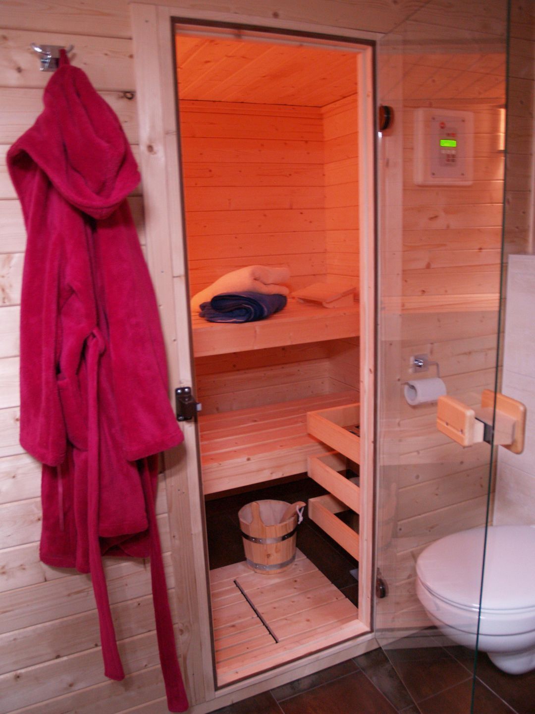 A wooden sauna room with a glass door, a red robe hanging on the wall, a wooden bench, a small wooden bucket, and a toilet.