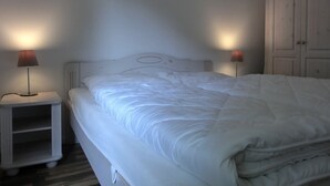 Una cama bien hecha con ropa de cama blanca, dos lámparas de noche y un armario blanco en una habitación sencilla.