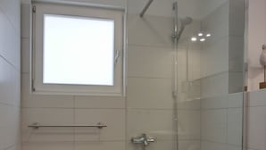 Un baño moderno con ducha de vidrio, inodoro blanco y un taburete pequeño.