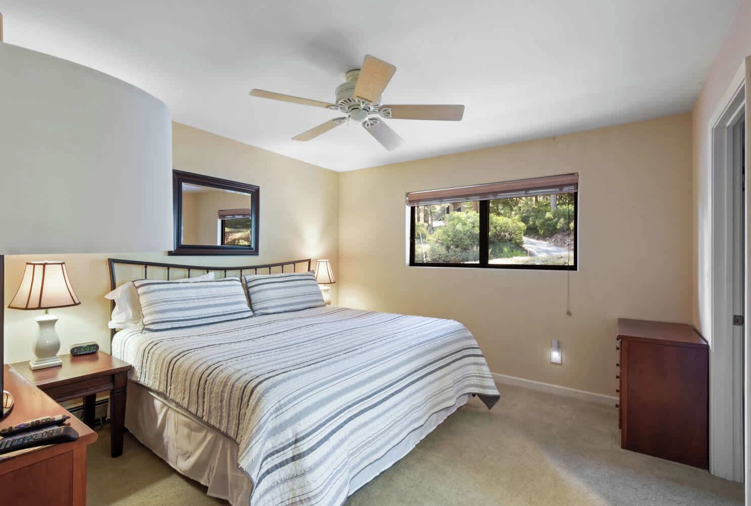 A bedroom with a bed, a nightstand, a lamp, a ceiling fan, a window, and a door.