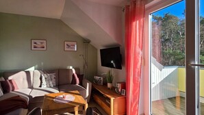 A cozy living room with a sofa, a coffee table, and a TV on a wall. There are two framed pictures on the wall, and a balcony with a red curtain.