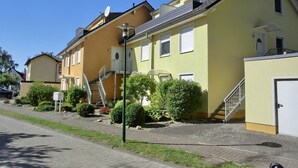 A yellow building with a staircase leading to a balcony, surrounded by greenery and a paved sidewalk.
