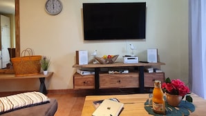 A living room with a flat-screen TV mounted on the wall, a wooden coffee table with a laptop, a bottle of juice, and a bowl of fruit.