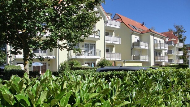 A multi-story residential building with balconies, a red-tiled roof, and a well-maintained hedge in the foreground.