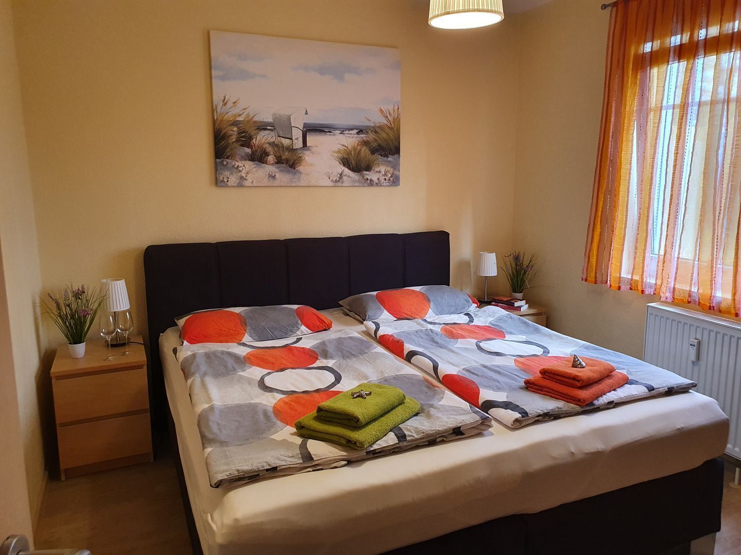 Un dormitorio con cama, mesita de noche, lámpara, ventana con cortinas y un cuadro en la pared.