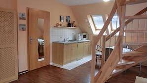 Una cucina compatta con mobili in legno, un lavello e un forno a microonde. C'è una zona pranzo con tavolo e sedie, e una scala in legno che conduce a un livello superiore.