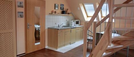 A compact kitchen with wooden cabinets, a sink, and a microwave. There is a dining area with a table and chairs, and a wooden staircase leading to an upper level.