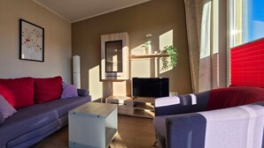 A modern living room with a sofa, a coffee table, and a TV. There is a window with curtains and a plant on the windowsill.