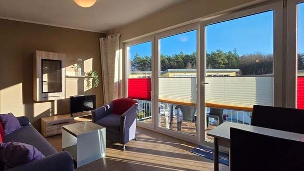 A modern living room with a sofa, coffee table, and TV, large windows with blinds, and a balcony with a view of a building and trees.