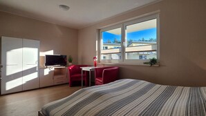 A bedroom with a bed, a TV, a desk, and a window with a view of buildings.