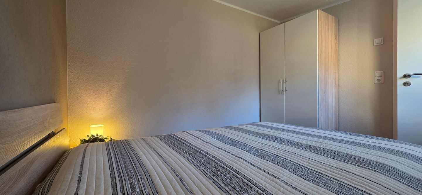 A bedroom with a bed, a nightstand, a wardrobe, and a wall-mounted light.