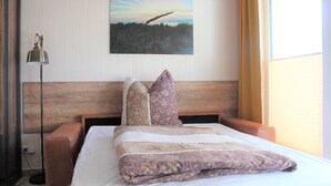 Ein ordentlich gemachtes Bett mit gemusterten Betttextilien, einem Holz-Kopfteil und einem Landschaftsbild an der Wand.