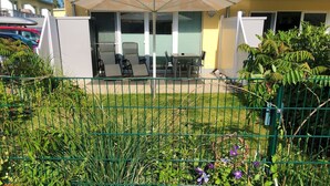Ein von einer Hecke umgebener Außenbereich mit Tisch und Stühlen unter einem Sonnensegel, umgeben von viel Grün und einem Gebäude mit gelbem Anstrich.