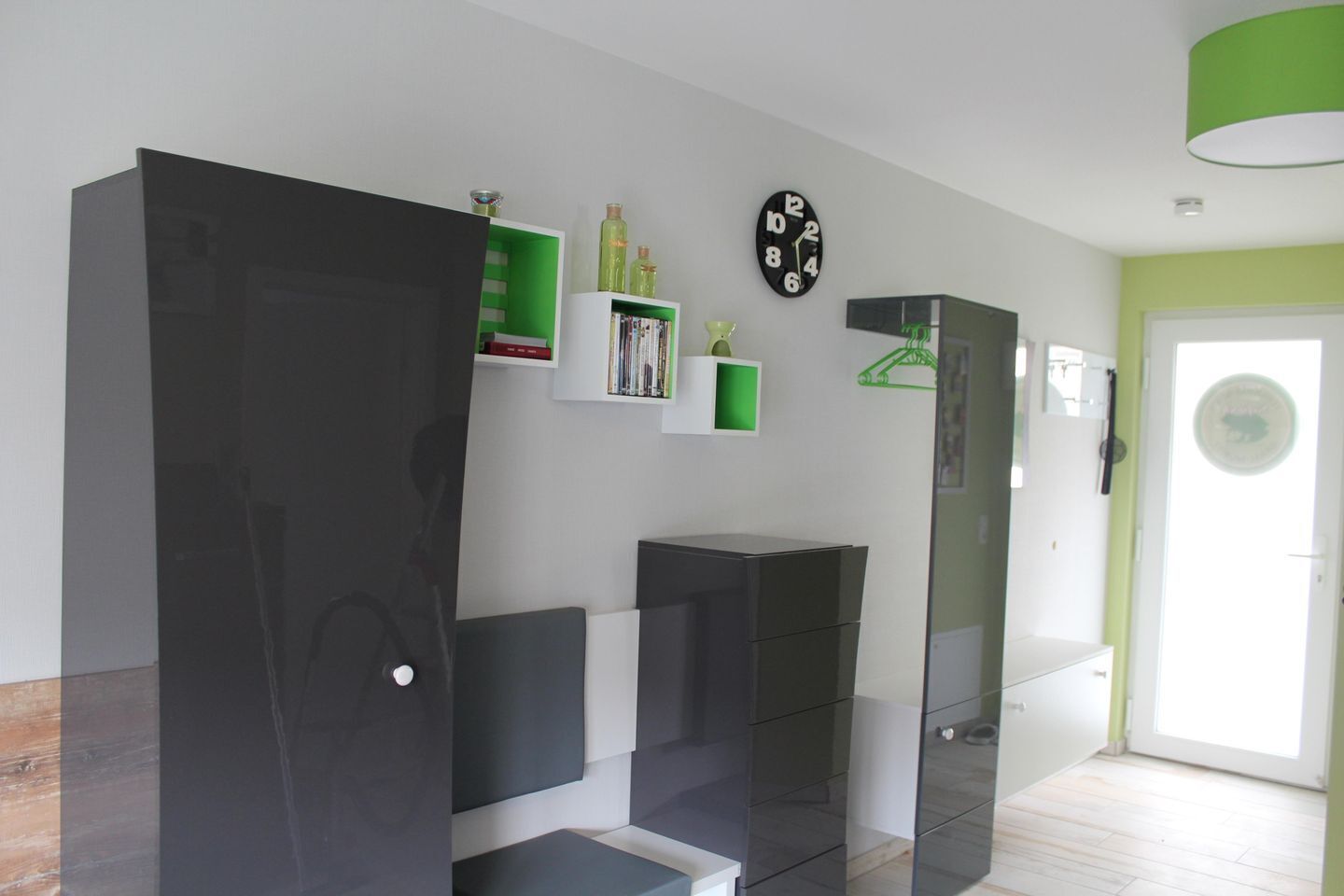 Un salon moderne avec un grand meuble noir, une étagère verte remplie de livres, une horloge noire et un luminaire suspendu vert.