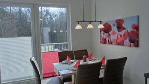 Un coin repas avec une table dressée pour quatre personnes, des chaises, une fenêtre avec des stores et un coussin rouge.