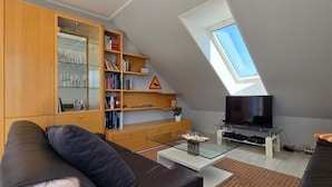 En moderne loftlejlighed med skråvægge, et sofabord i glas, en sofa, et fjernsyn på et glasbord og en reol med diverse genstande.