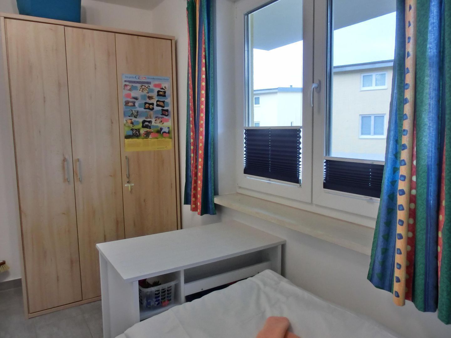 A bedroom with a wooden wardrobe, a white desk, a bed with a white mattress, and a window with blinds.