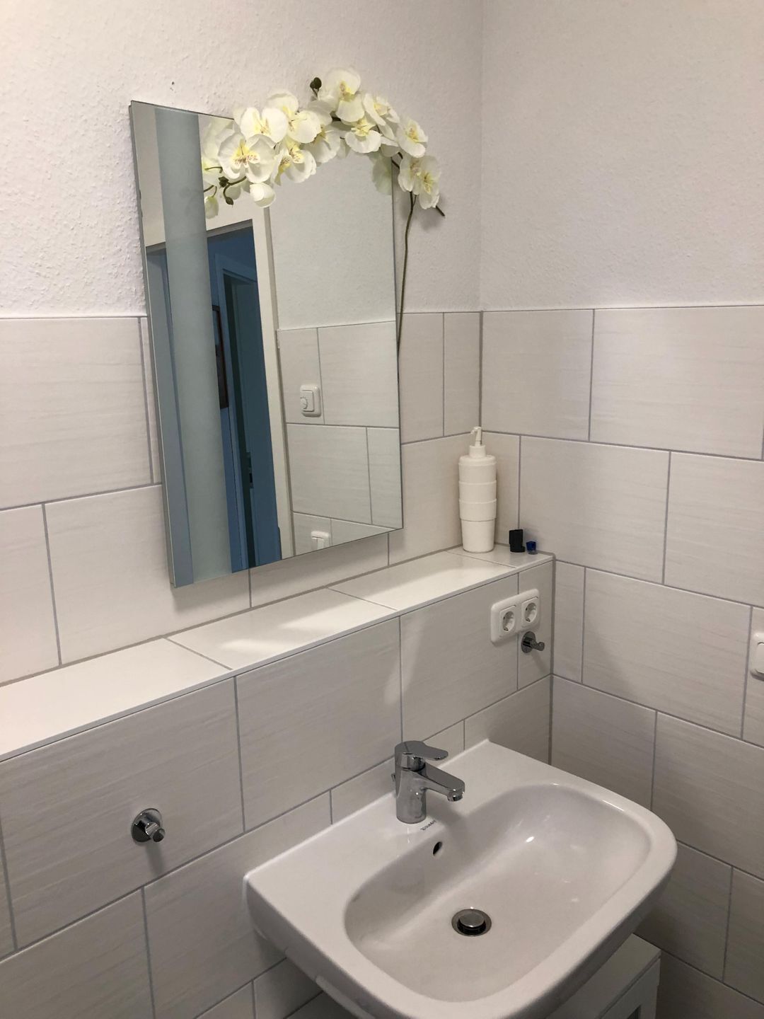 A bathroom with a white sink, a mirror, and a white flower arrangement.