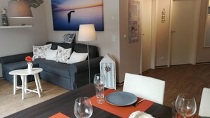 A living room with a grey sofa, a white table with a vase of flowers, and a painting of a sunset.