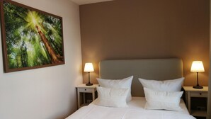 A neatly made bed with white bedding and pillows, two bedside tables with lamps, and a large framed picture of a forest on the wall.