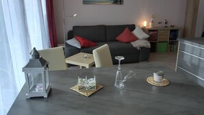 Ett modernt vardagsrum med en grå soffa, röda och vita kuddar, en tavla som föreställer en solnedgång, en golvlampa, ett litet bord med ett ljus, en glasvas och en dekorativ lykta.