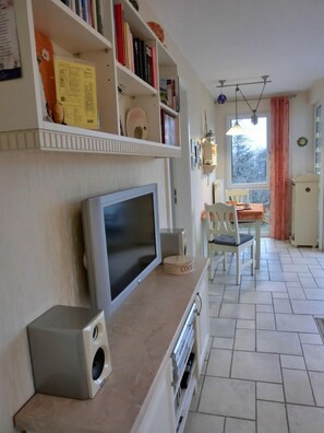 A living room with a television, bookshelves, a dining area with a table and chairs, and a kitchen area with a refrigerator.
