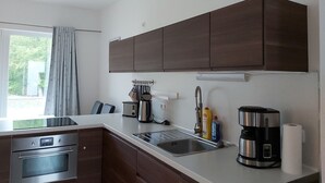 A modern kitchen with dark wooden cabinets, stainless steel appliances, a built-in oven, a coffee maker, and a window with a view of greenery. - Apartment KWi 26 OG - 2-room apartment for 3 pers. WLAN (Gelbensande)