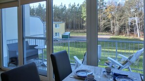 A dining area with a table set for two, overlooking a grassy area with trees and a building in the background. - Apartment KWi 26 OG - 2-room apartment for 3 pers. WLAN (Gelbensande)