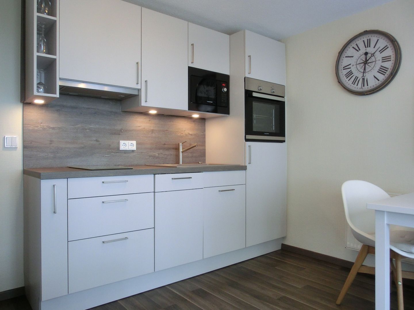 A modern kitchen with white cabinets, a built-in oven, a microwave, and a clock on the wall.