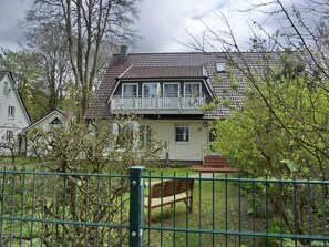 Une maison à deux étages avec un balcon, entourée d’arbres et d’une clôture.