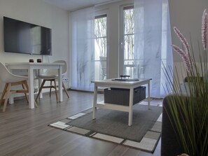 Un salon moderne avec un téléviseur à écran plat, un bureau blanc, deux chaises, une petite table avec un panier, et une grande fenêtre avec des stores.