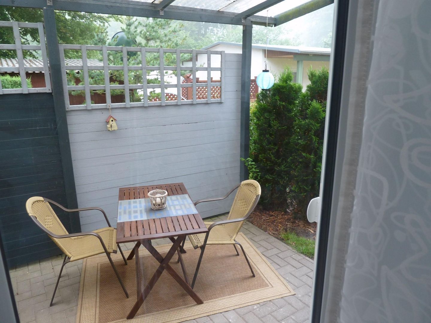 An outdoor patio with a wooden table and two wicker chairs, a white fence, and a house in the background.