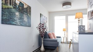 A modern living room with a grey sofa, a vase with dried flowers, and a painting of a cityscape. - Apartment KWi 83 ground floor "Casa Baltico" - 2-room apartment for 3 pers. with WLAN (Gelbensande)