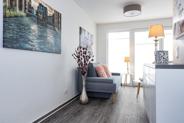 A modern living room with a grey sofa, a vase with dried flowers, and a painting of a cityscape.