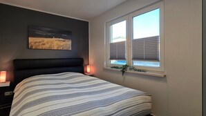 Ein Schlafzimmer mit einem Bett, einem Nachttisch mit Lampe, einem Fenster mit Jalousien und einem Wandbild.