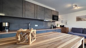 Eine moderne Küche mit dunklen Schränken, einer Mikrowelle und einer Kaffeemaschine. Es gibt einen Holztisch mit einer Tischdekoration aus Seil. Im Wohnbereich befinden sich ein blaues Sofa und eine kleine Holzbank.