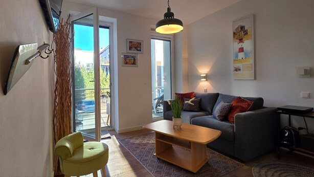 A modern living room with a sofa, a coffee table, a chair, and a large window. - Vacation apartment LP14 - 1-room vacation apartment with approx. 19 m² living space and balcony (Graal-Müritz)