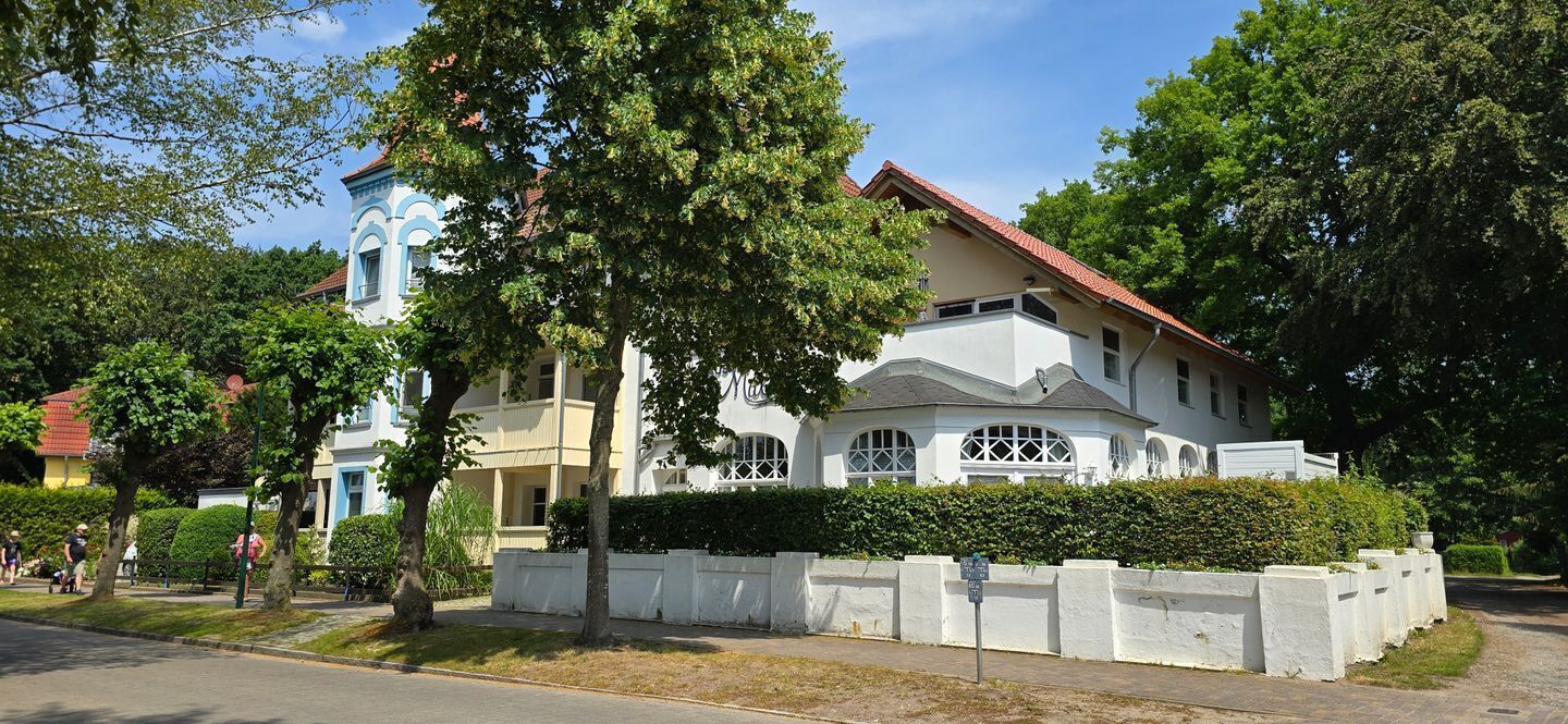 A large, two-story building with a white facade and red roof, surrounded by lush greenery and trees.