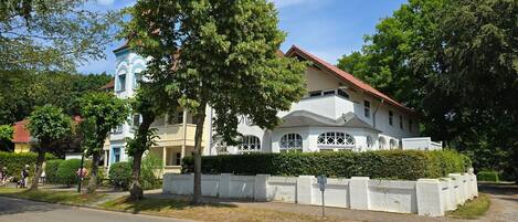 A large, two-story building with a white facade and red roof, surrounded by lush greenery and trees.