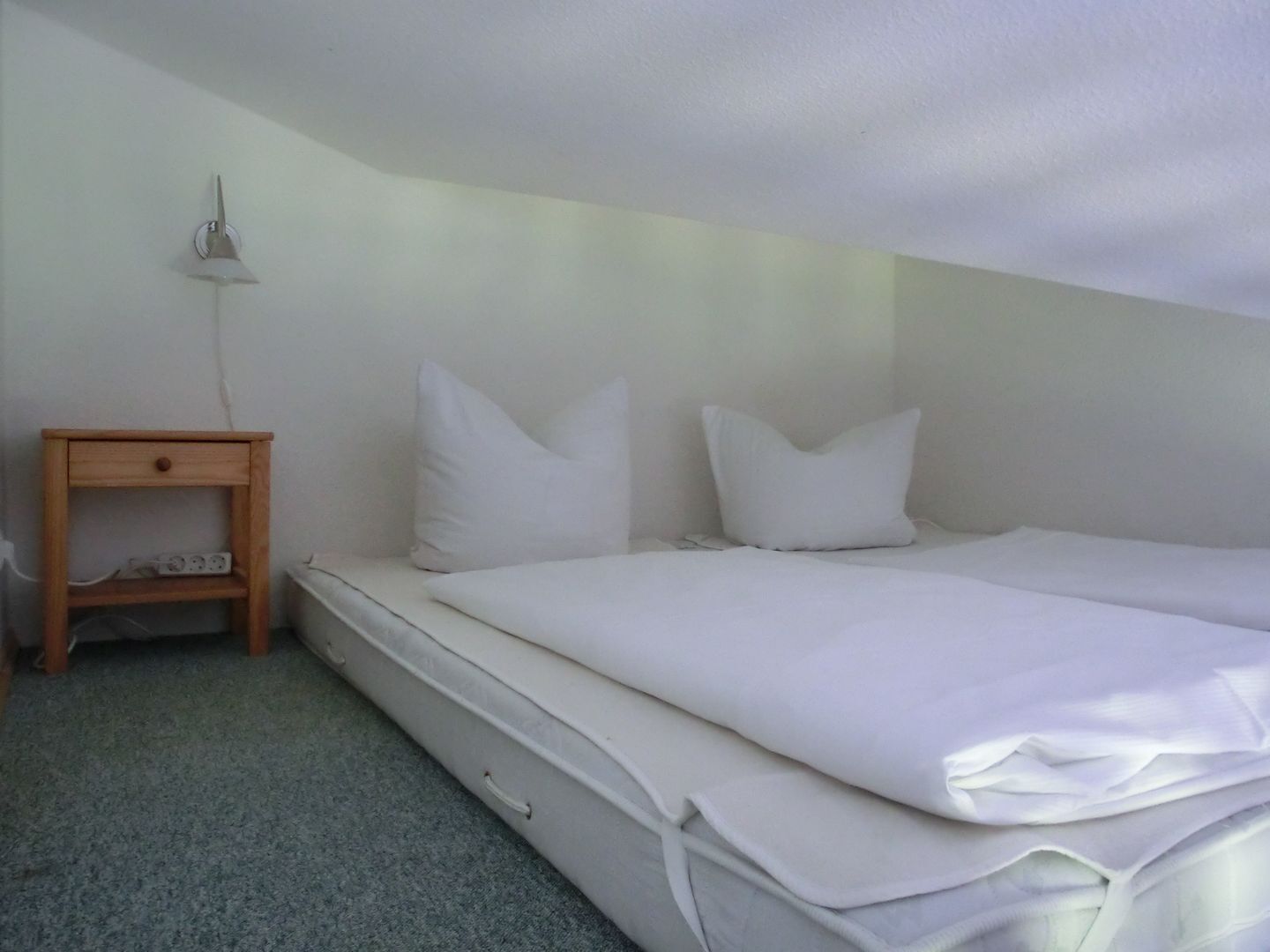 Un lit simple avec du linge de lit blanc, une petite table de chevet en bois avec une lampe et une prise électrique.