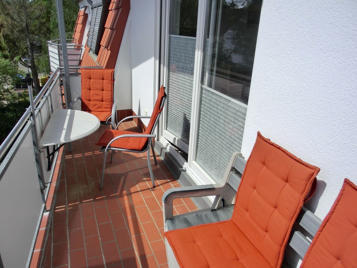 A balcony with orange chairs and a small table, a white wall, and a window with blinds.