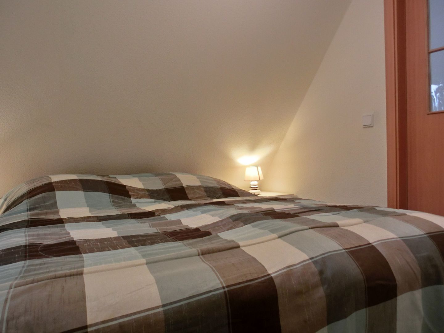 Een slaapkamer met een geruit beddegoed, een bedlampje en een houten deur.