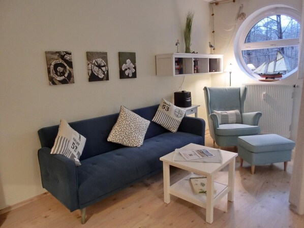 A living room with a dark blue sofa, a light blue armchair, a white coffee table, and a round window with a view of trees.