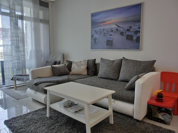 A modern living room with a grey sofa, white coffee table, and a red chair. A painting of a beach scene hangs on the wall.