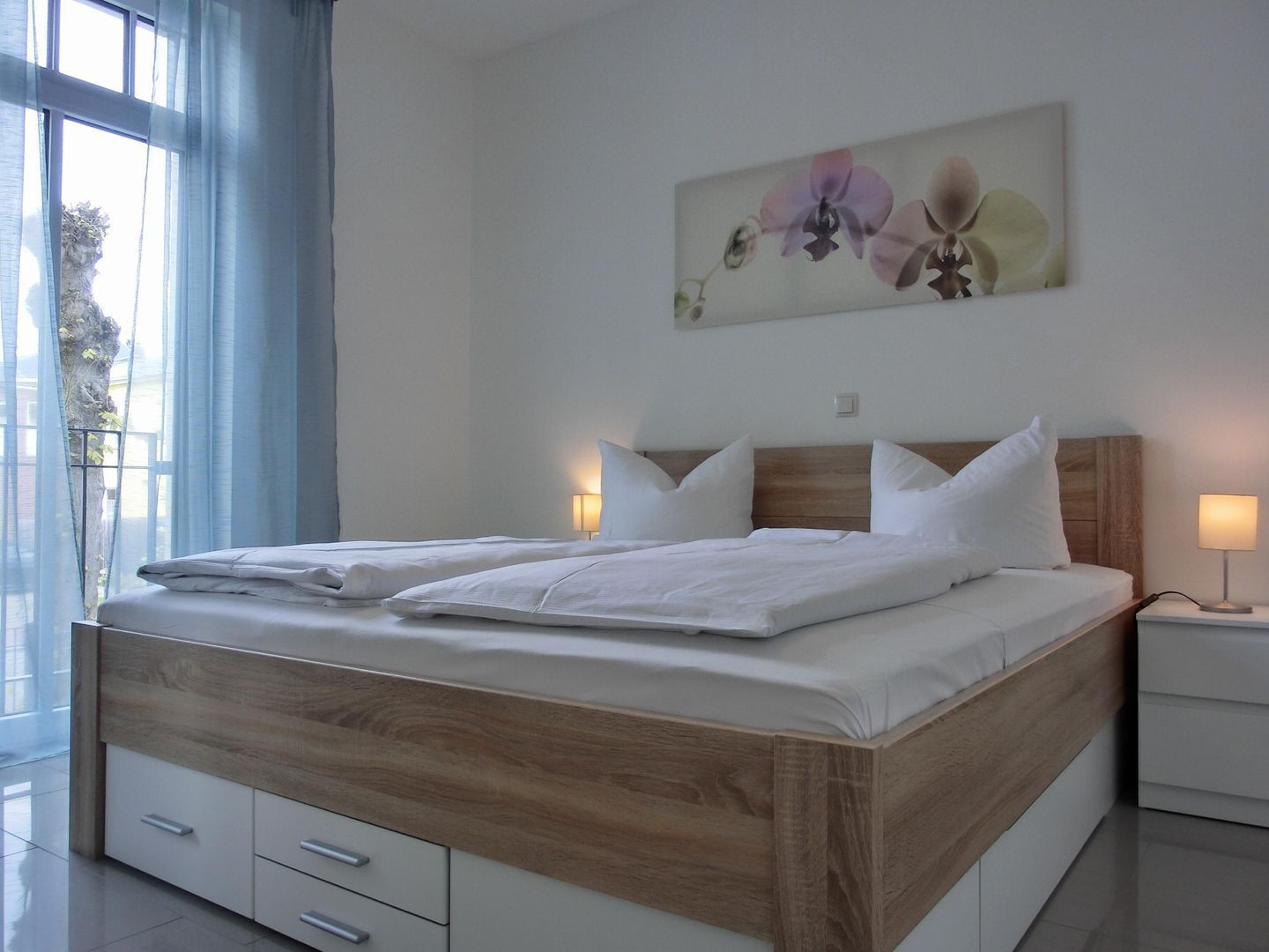 Ein modernes Schlafzimmer mit einem Holzbettgestell, weißer Bettwäsche und einem großen Fenster mit blauen Vorhängen.