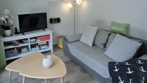 Una sala de estar moderna con un televisor de pantalla plana, estanterías, un sofá gris con cojines estampados, una pequeña mesa de madera y una lámpara de pared.